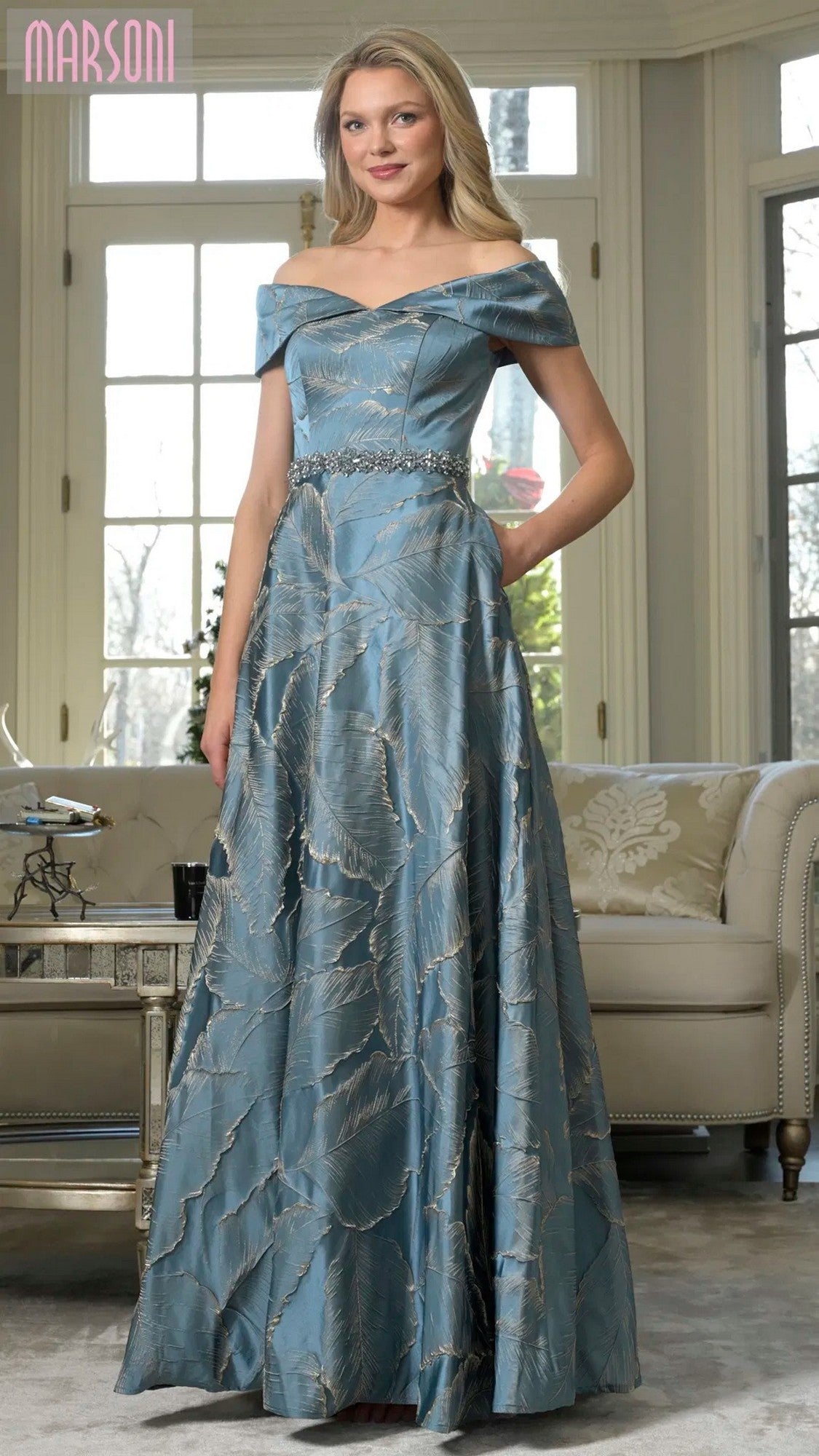 A woman stands indoors wearing the Marsoni MV1265 long formal dress in teal, featuring an off-the-shoulder design, leaf pattern, and sparkling beaded waistline for an elegant look.